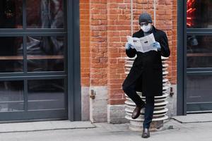 Ganzkörperaufnahme eines ernsthaften Mannes in schwarzem Mantel, trägt eine Sonnenbrille und eine medizinische Maske, liest Zeitung, posiert gegen eine Ziegelwand und schützt sich vor Coronavirus. Epidemie und Quarantäne foto