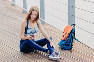 schöne ernsthafte frau hat einen blick auf den bildschirm des smartphones, prüft, welches lied sie hören soll, verwendet moderne kopfhörer zur unterhaltung, sitzt mit gekreuzten beinen in der nähe der tasche im freien, ruht sich nach dem training aus. foto