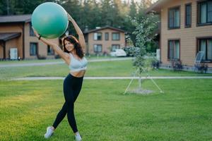 sportlich schlanke frau trägt bauchfreies top und leggings, macht aerobic-übungen mit fitnessball, posiert im freien auf grünem rasen, privathäuser im hintergrund. gesunde fit weibliches model hebt fitball über den kopf foto