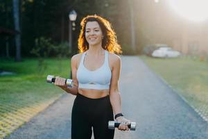 Foto einer schlanken, brünetten jungen Frau, die Hanteln hebt, morgendliche Trainingsposen gegen Sonnenaufgang hat, in einem kurzen Oberteil und Leggings gekleidet ist, arbeitet an den Armmuskeln, lächelt positiv und führt zu einem sportlichen Lebensstil