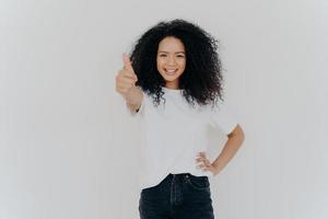 Foto einer glücklichen lockigen Frau mit knackigem Haar hebt Daumen hoch, gibt Zustimmung, sagt, klingt gut, macht unterstützende Geste, hält die andere Hand auf der Taille, lässig gekleidet, isoliert auf weißem Hintergrund