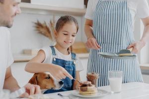 Fröhliches kleines Mädchen isst gerne leckeres Dessert, das von Mama zubereitet wird, fügt Pfannkuchen geschmolzene Schokolade hinzu, genießt das Zusammensein und Mutter, Vater und Hund genießen ein köstliches, nahrhaftes Frühstück in der Küche foto