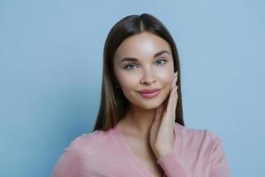 Foto einer entzückenden jungen tausendjährigen Frau berührt sanft die Wange, genießt die Weichheit der Haut nach dem Auftragen einer Schönheitsmaske oder Gesichtscreme, blickt in die Kamera, isoliert auf blauem Hintergrund. Weiblichkeitskonzept