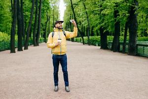 Horizontales Porträt eines gutaussehenden bärtigen Mannes in gelbem Anorak, Mütze und Jeans mit glücklichem Gesichtsausdruck, während er im Park posiert und Selfie macht, während er Kaffee trinkt. Hipster, der von Selfie begeistert ist foto