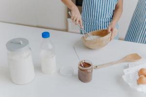 7/8-Ansicht von Kind in Schürze verquirlt Zutaten in Schüssel mit Rührbesen, eifrig kochen und Mutter helfen, Kuchen zuzubereiten. teig, milch, eier, holzspatel und schokolade auf dem tisch foto