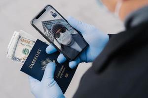 ein nicht erkennbarer mann hat einen videoanruf, spricht mit einem freund, trägt schützende medizinische gummihandschuhe, hält einen reisepass mit geld und wird das land während einer coronavirus-pandemie verlassen. Konzept der sozialen Distanz foto