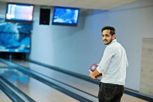 südasiatischer mann im jeanshemd, der an der kegelbahn mit ball auf händen steht. Kerl bereitet sich auf einen Wurf vor. foto