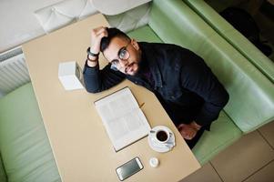 arabischer mann trägt schwarze jeansjacke und brille, sitzt im café, liest buch und trinkt kaffee. stilvoller und modischer arabischer Modelltyp. foto