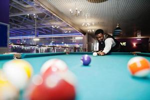 Junger gutaussehender afrikanischer Mann mit weißem Hemd, schwarzer Weste und Fliege spielt Billard. foto