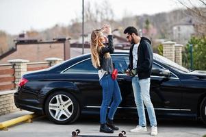 cooles gemischtrassiges Paar umarmt sich zusammen mit Longboard gegen schwarzen eleganten Sportwagen. foto