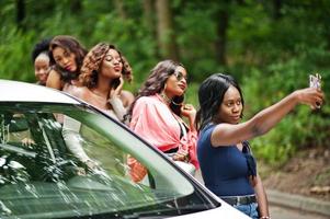 gruppe von fünf glücklichen afroamerikanischen reisenden mädchen, die selfie gegen auto machen. foto