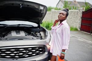 junge traurige afrikanerin steht mit notdreieck gegen auto mit geknallter motorhaube. Transport, Fahrzeugprobleme und Pannenkonzept. foto