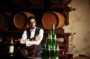 hübscher, gut gekleideter arabischer mann mit einem glas whisky und einer zigarre, die im pub posiert. foto
