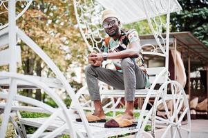 gutaussehender afroamerikanischer mann mit traditioneller kleidung, mütze und brille sitzt auf einem weißen wagen. foto