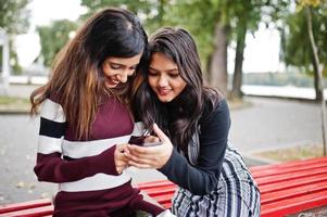 Porträt von zwei jungen schönen indischen oder südasiatischen Mädchen im Teenageralter im Kleid, die auf einer Bank sitzen und ein Handy benutzen. foto