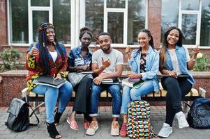 Gruppe von fünf afrikanischen College-Studenten, die gemeinsam Zeit auf dem Campus des Universitätshofs verbringen. schwarze afrofreunde, die an der bank mit schulsachen, laptops notizbüchern studieren. foto