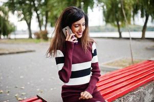 Porträt eines jungen, schönen indischen oder südasiatischen Teenager-Mädchens im Kleid, das mit einem Handy auf einer Bank sitzt. foto