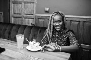 porträt einer schönen jungen afrikanischen geschäftsfrau mit dreadlocks, blauer bluse und rock, die im café mit eis und ananassaft sitzt. sie schaut auf uhren an ihrer hand. foto