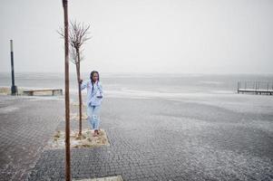 Stilvolles afrikanisch-amerikanisches Mädchen mit Dreadlocks, das Handy zur Hand hält, im Freien auf Pier gegen zugefrorenen See bei Schneewetter. foto