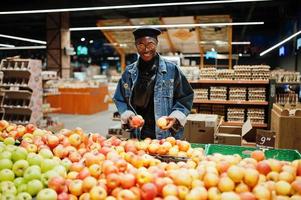 Stilvoller lässiger afroamerikanischer Mann in Jeansjacke und schwarzer Baskenmütze, der Apfelfrüchte in der Bio-Abteilung des Supermarkts überprüft. foto