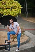 modischer großer arabischer bartmann trägt hemd, jeans und sonnenbrille mit regenschirm, der bei regen auf dem parkplatz posiert. foto