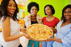 Vier junge afrikanische Mädchen in einem farbenfrohen Restaurant halten ein Holztablett mit Pizza in den Händen. foto