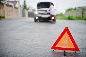 junge traurige afrikanerin steht mit notdreieck gegen auto mit geknallter motorhaube. Transport, Fahrzeugprobleme und Pannenkonzept. foto