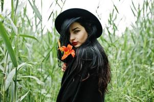 sinnliches Mädchen ganz in Schwarz, roten Lippen und Hut. gotische dramatische frau hält orangefarbene lilienblume auf gemeinem schilf. foto