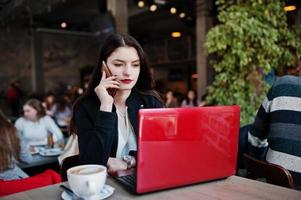 Brünettes Mädchen sitzt im Café, arbeitet mit rotem Laptop und spricht am Handy. foto