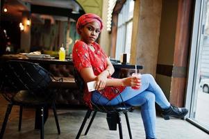 stilvolle afrikanische frau in rotem hemd und hut posierte innencafé und trank erdbeerlimonade mit telefon zur hand. foto