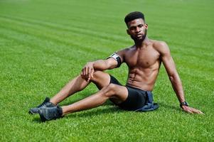 afroamerikanischer männlicher athlet sexy sport nackter oberkörper mann mit laufendem sportarmgehäuse für handy, posiert auf grünem gras des fußballstadions. foto