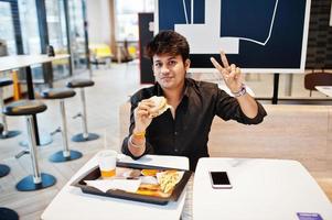stilvoller indischer mann, der im fast-food-café sitzt und hamburger isst und friedenszeichenhand gestikuliert. foto