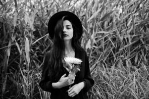 sinnliches Mädchen ganz in Schwarz, roten Lippen und Hut. gotische dramatische frau auf gemeinem schilf halten weiße chrysanthemenblume. Schwarz-Weiß-Porträt. foto