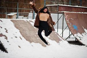 Afroamerikanische Frau in Schaffellmantel und Mütze posiert und springt am Wintertag vor schneebedecktem Hintergrund. foto