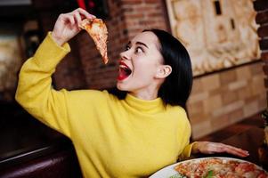 lustiges brünettes mädchen im gelben pullover, der pizza im restaurant isst. foto