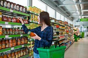 Einkaufsfrau, die die Regale im Supermarkt betrachtet. Porträt eines jungen Mädchens in einem Marktladen mit grünem Einkaufskorb und einer Dose Gemüse. foto