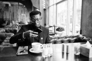 modischer afroamerikanischer mann in anzug und brille sitzt im café und schnappt kaffee am telefon. foto