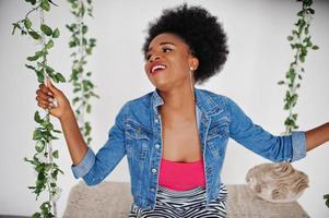 Attraktive Afroamerikanerin mit Afro-Haar auf Rock und Jeansjacke, posiert im weißen Raum auf Schaukel. modisches schwarzes Modell. foto