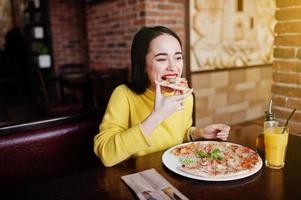 lustiges brünettes mädchen im gelben pullover, der pizza im restaurant isst. foto