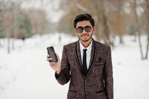 stylischer indischer studentenmann im braunen anzug und sonnenbrille posierte am wintertag im freien mit handy zur hand. foto