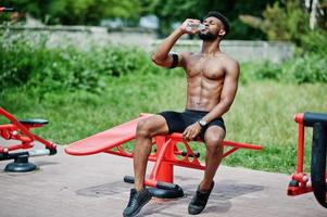 afroamerikanischer männlicher athlet sexy sport mit nacktem torso mann mit laufendem sportarmgehäuse für handy, übungen auf den straßentrainingssimulatoren und trinkwasser aus der flasche. foto