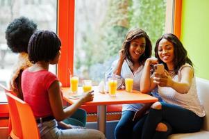 vier junge afrikanische mädchen mit säften sitzen in einem bunten fast-food-restaurant und machen selfie per telefon. foto