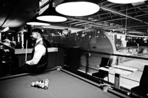 Junger gutaussehender afrikanischer Mann mit weißem Hemd, schwarzer Weste und Fliege spielt Billard. foto