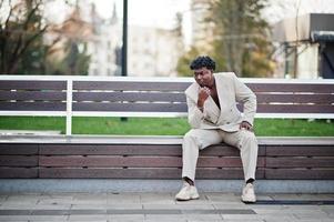 Stilvoller Afro-Mann im beigefarbenen Anzug der alten Schule sitzt auf der Bank. Modischer junger afrikanischer Mann in Freizeitjacke auf nacktem Oberkörper. foto