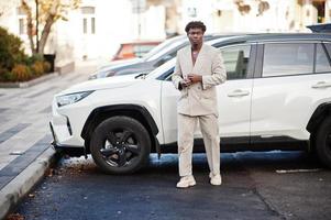 Stylischer Afro-Mann in beigem Old-School-Anzug gegen modernes Hybrid-Elektro-SUV-Auto. Modischer junger afrikanischer Mann in Freizeitjacke auf nacktem Oberkörper. foto