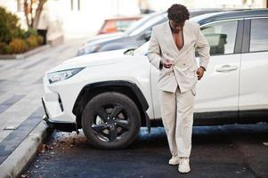 Stylischer Afro-Mann in beigem Old-School-Anzug gegen modernes Hybrid-Elektro-SUV-Auto. Modischer junger afrikanischer Mann in Freizeitjacke auf nacktem Oberkörper. foto