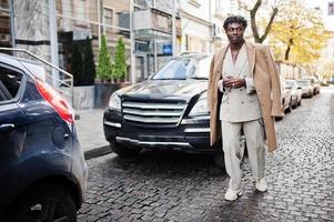 Stilvoller Afro-Mann in beigefarbenem Anzug und Mantel der alten Schule, der gegen ein schwarzes Geschäftsauto läuft. Modischer junger afrikanischer Mann in Freizeitjacke auf nacktem Oberkörper. foto