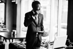 business afroamerikaner trägt schwarzen anzug und brille im büro mit laptop und trinkt morgens kaffee vor dem arbeitstag. foto