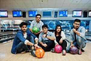 gruppe von fünf südasiatischen völkern, die sich im bowlingclub ausruhen und spaß haben und mit bällen auf den händen auf der bowlingbahn sitzen. foto