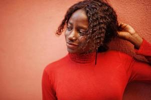 Porträt einer lockigen Afrikanerin mit modischem rotem Rollkragenpullover, die an der Rosenwand posiert. foto
